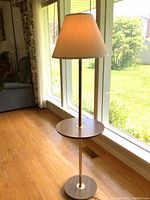 Full view of vintage brass floor lamp with integrated wooden table and beige pleated fabric shade lit by natural window light, showing height and style.