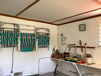 Wide angle view showing three vintage woven folding garden chairs mounted on the wall and various gardening tools and accessories on a wall shelf beneath a green framed wall clock.