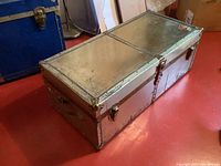 Top angled view of the large rectangular metal trunk on a red floor, showing metal edges, side handle, and hinged lid divided in two panels with brass clasp hardware.