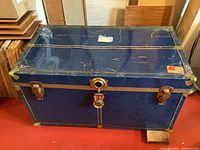 Front view of large blue metal-covered wooden storage trunk showing the overall shape, size, and condition with visible scratch marks and metal corner reinforcements.