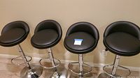 Four black cushioned adjustable swivel bar stools with metal pedestal bases and footrests, aligned against a wall on a wooden floor. One stool has a bent backrest.