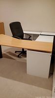 Right angled two tone desk shown with birch desktop, white cabinets and black swivel office chair.