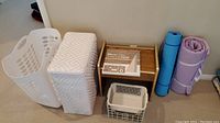 Photo showing plastic and wooden laundry hampers, two rolled yoga mats, a small white plastic magazine container, and boxed mirror tiles on floor.