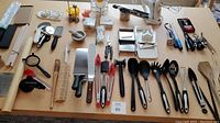 Full overview of kitchen gadgets including spatulas, tongs, graters, peelers, chopsticks, whisks, mortar and pestle, pizza cutter, and more spread on table.