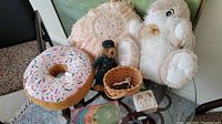 Overview photo showing stuffed bunny plush, donut cushion, teddy bear toy, small basket, and angel figurines on glass table