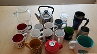 Overall view of a collection of mugs of different sizes and designs plus a stainless steel tea kettle and clear glass pitcher on a light wood table.