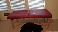 Full side view of Oakworks burgundy colored massage table with wooden legs and folded black carrying case below.