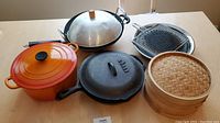 Overview of cast iron cookware including Le Creuset pot, cast iron skillets with one lid, wok, and grilling racks with bamboo steamer set.