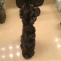 Statue showing two African women, one standing and one seated, with beaded necklaces and textured clothing, placed on a reflective surface with overhead lighting.