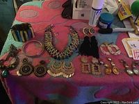Photo showing multiple pieces of costume jewelry arranged on a colorful cloth, including a chunky purple stone necklace, large earrings, tassel earrings, bangles, and bracelets.