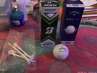 Photo showing Callaway E.R.C. Soft and Bridgestone RXS golf ball boxes with a Callaway golf ball and pack of white golf tees on a cloth surface.