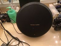 Front view of round black Harman Kardon speaker placed on counter with attached power cord and power adapter visible.