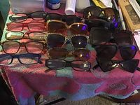 Top-down photo showing 15 pairs of eyewear arranged on a towel, including sunglasses and reading glasses in various frame colors and styles.