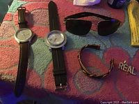Photo showing two men's wristwatches, one pair of black aviator sunglasses, a twisted metal bracelet, and a rhinestone 'REAL' pin all laid out on a multi-colored towel.