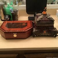Two decorative boxes side by side on countertop, showing wooden box with glossy reddish-brown finish and ornate box with detailed leaf carvings and amber knob on top, lid detached.