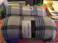 Two rolled Mainstays fleece throws with plaid pattern in gray, cream, and blue on a red surface.