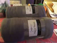 Two rolled gray fleece throws with Mainstays label, placed on a table with other items in background.