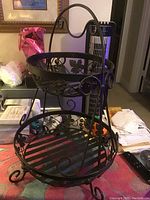 Photo of vintage two-tier metal fruit or vegetable holder with scrollwork and leaf decorative details, placed on a table with background household items.