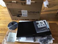 Monitor partially unpacked showing Asus VP249 branding on the back, its round black stand base, cables, manuals, and protective plastic still on the screen.