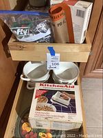 Wooden box with boxed KitchenAid mixer, plastic bag of cookie cutters, silicone mat, and four white bowls below