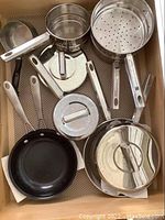 Top-down view of various stainless steel cookware items including a steamer insert, pots with lids, and a non-stick fry pan all inside a drawer.