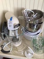 Front view of mixer with glass bowl, stainless steel bowls, plastic bowl, measuring cups, attachments on countertop