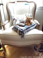 Front view of cream brocade armchair with decorative items on the seat including teddy bears, swan plush, pin cushion, and blanket.