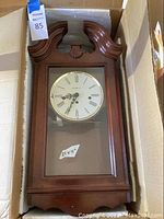 Howard Miller wall clock in original box, wooden case with glass front, clock face with Roman numerals, labeled dimensions 21x11 inches.