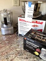 KitchenAid Food Grinder attachment in box beside Cuisinart food processor and mixing bowl on countertop.
