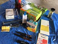 Photo of entire lot showing all items together on blue tarp, including Swiffer box, carbon monoxide alarm box, flashlights, utensils, and brass lantern