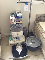 Stacked bathroom paper goods with toilet paper rolls, facial tissue boxes, and plastic wrapped packages near the corner. Adjacent to these items is a silver metal towel rack and a gray commode bucket with lid. Mechanical bathroom scale is in front of the paper goods.