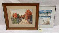 Two framed watercolor paintings side by side, one autumn scene with orange and red trees along a dirt road, one winter scene with snow and bare trees.