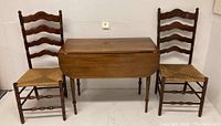 Front view of the vintage drop leaf table between two ladder back chairs with rush seats showing overall condition and size.
