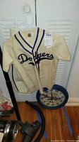 Child's Brooklyn Dodgers baseball jersey hung on a white plastic hanger showing front with large Dodgers script logo, blue collar piping, with matching pants behind and Dodgers wall clock on floor.