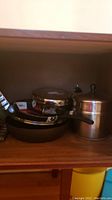 Photo of metal pots and pans including a non-stick frying pan with handle, a medium-size pot with lid and another small metal pot or container with lid.