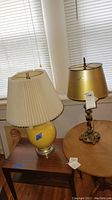 Two table lamps by a window on a wooden table, one with a yellow round base and pleated shade, the other ornate gold metal base with gold shade.