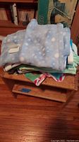 Wooden shelf with several small folded blankets including a blue polka dot fleece blanket on top, set on wooden floor with various household surroundings.