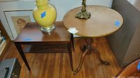 Photo showing both side tables side by side on a wood floor with a yellow lamp and framed art leaning behind.