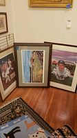 Three baseball themed framed art pieces leaning against a wall on a wooden floor with a patterned rug nearby. Includes Ebbet's Field print and player portrait.