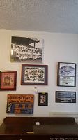 Seven baseball themed wall art pieces hanging on a wall above a wooden piece of furniture. The artwork includes black and white photos, colorful illustrations, and printed memorabilia.