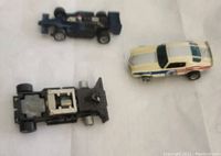 Three slot cars displayed on a sheet, including white Camaro with stripes, dark blue chassis only, and another chassis without body