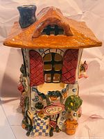 Front view of the Blue Sky Clayworks ceramic house figurine showing front door with 'Cafe' sign, windows with shutters, flower arrangements, and orange roof with chimney.