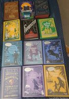 Top view of 12 hardcover books arranged in three rows on a fabric surface. Shows titles and cover designs, some still sealed in plastic.