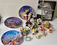 Image showing a group of Disney collectibles including three decorative plates with images of castles and Disney characters, various character figurines such as Mickey Mouse, Minnie Mouse, Dumbo, Tinker Bell, and others, along with a decorative item with a silver ribbon.