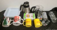 Wide view of mugs including Starbucks, Pirelli Tires, and mid-century style glasses along with a stainless steel coffee carafe and packaged creamers.