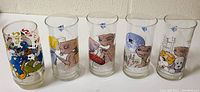 Five clear cylindrical drinking glasses showing colorful 1980s Smurfs and E.T. designs including E.T. characters and cartoon Smurfs action poses.