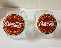 Pair of clear glass candy jars with red and yellow Coca-Cola lids showing the top logo design.
