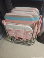 Four folded lawn chairs stacked together showing stripes of pink, blue, tan, and white on fabric seats and backs with metal frames.