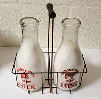 Front view of both vintage glass milk bottles sitting inside the metal wire caddy showing the Hickory Hills Dairy milk and buttermilk graphics.