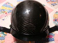 Front view of glossy black carbon Kevlar helmet with Harley Davidson logo on top, showing weave pattern and wear on ear covers.